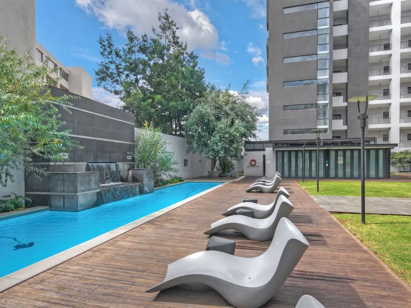 A well-maintained communal pool area featuring a modern water feature, timber decking, and landscaped garden space within an apartment complex.