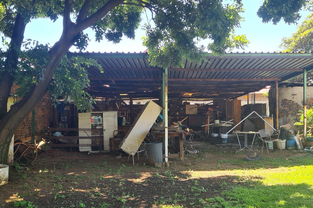 A rear outdoor area featuring a large corrugated metal roof structure supported by steel columns, set within a garden space with mature trees.