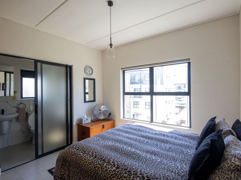 A well-lit bedroom featuring a modern en-suite bathroom with a sliding glass door, neutral finishes, and large windows providing natural light.