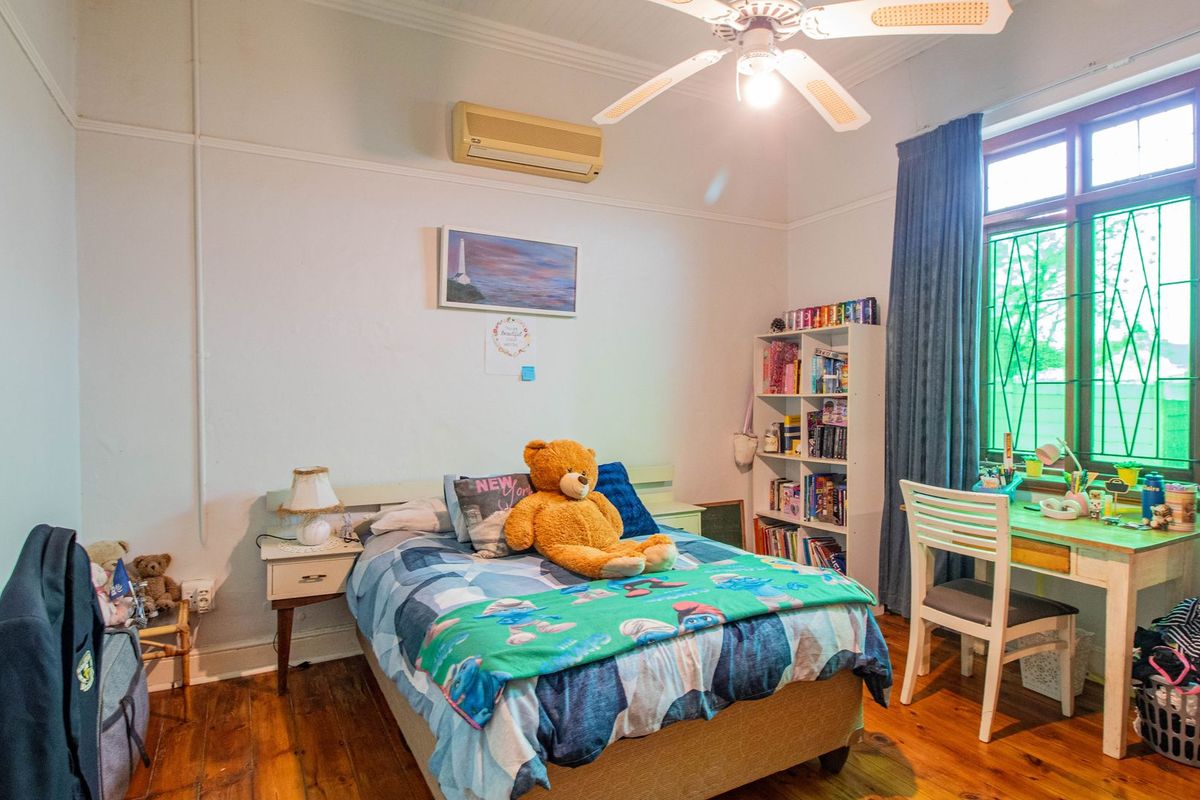 A bedroom featuring polished timber flooring, a ceiling fan, and a wall-mounted air conditioning unit, with large windows providing natural light.