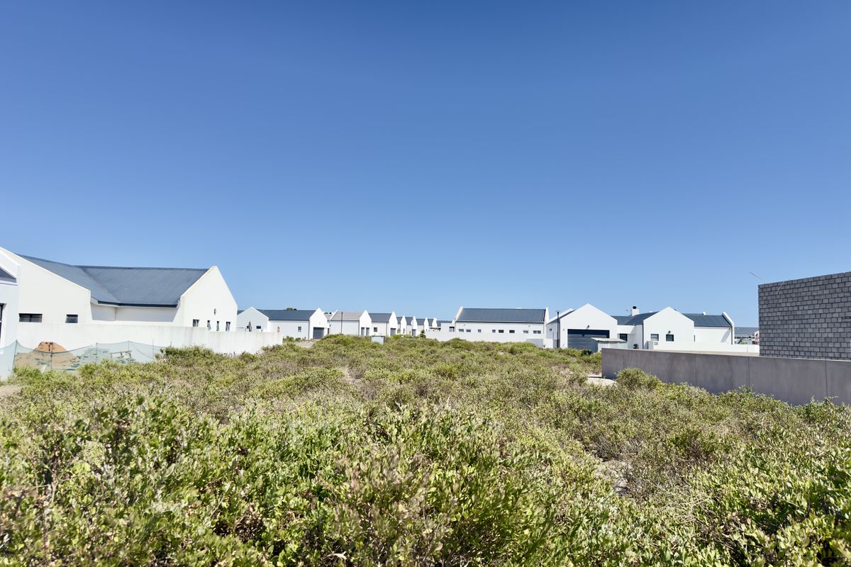 A view of a vacant residential plot covered in natural coastal vegetation, situated within a developed estate with surrounding white-walled homes.