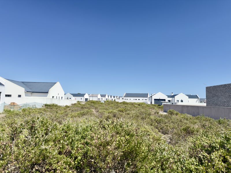 A view of a vacant residential plot covered in natural coastal vegetation, situated within a developed estate with surrounding white-walled homes.