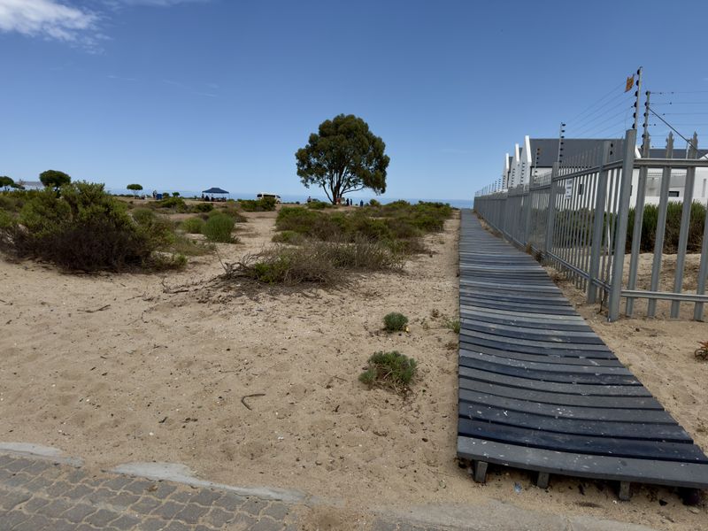 A sandy coastal residential plot featuring a timber boardwalk path alongside a metal perimeter fence with electric security wiring.