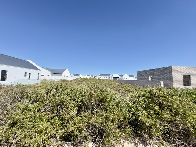 A view of a vacant residential plot featuring native coastal vegetation, adjacent to existing residential developments with established boundary walls.