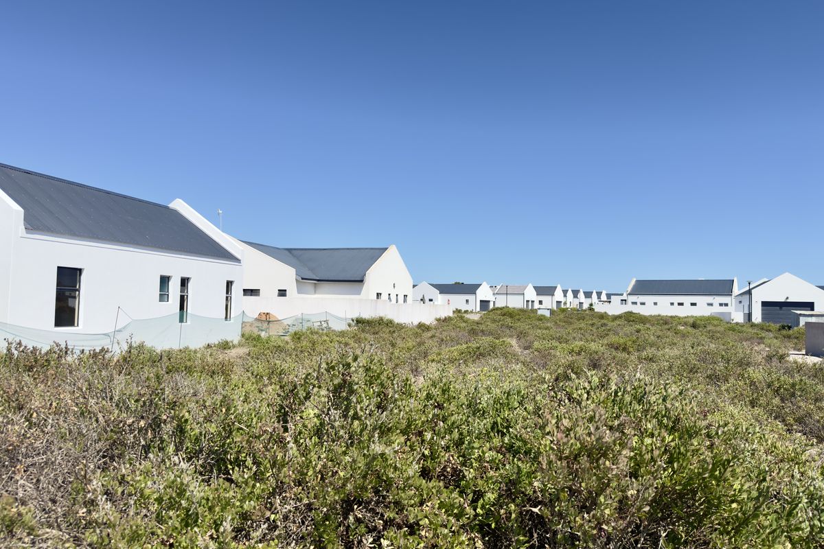 A view of a vacant residential plot featuring natural coastal vegetation, with established modern white homes visible in the background.