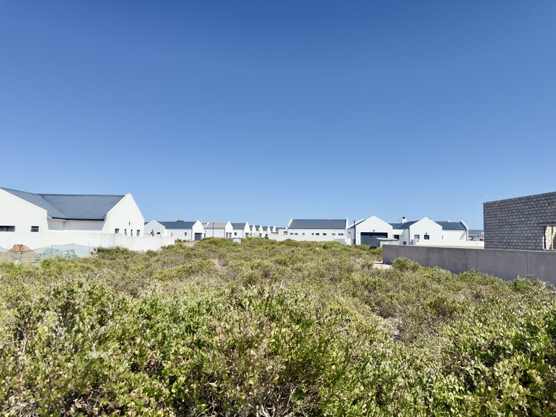 A vacant residential plot covered in native coastal vegetation, situated within a developed estate with surrounding modern residential properties.