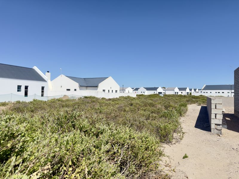 A view of a vacant residential plot featuring natural vegetation, adjacent to established housing developments in a coastal or estate setting.