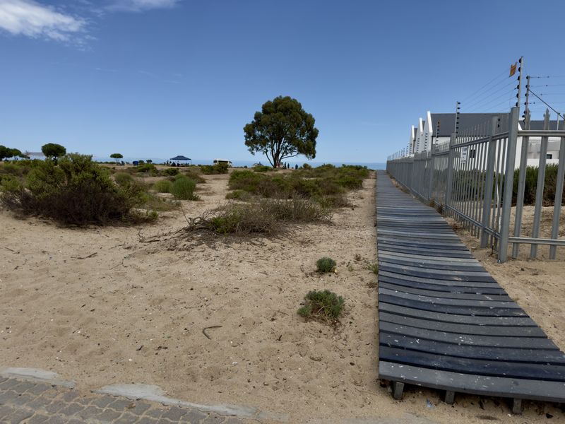 A sandy coastal plot featuring a timber boardwalk path alongside a fenced residential property boundary, leading towards the ocean.