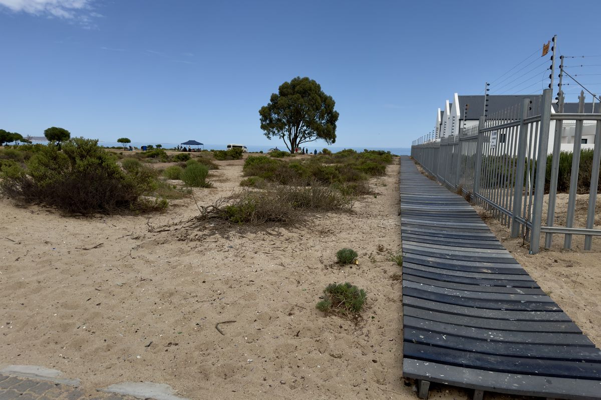 A sandy coastal plot featuring a timber boardwalk path alongside a fenced residential property boundary, leading towards the ocean.