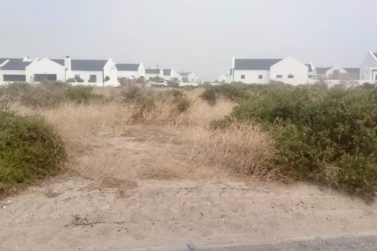 A vacant residential plot covered in natural coastal vegetation, situated within a developed estate with established white-walled homes in the background.