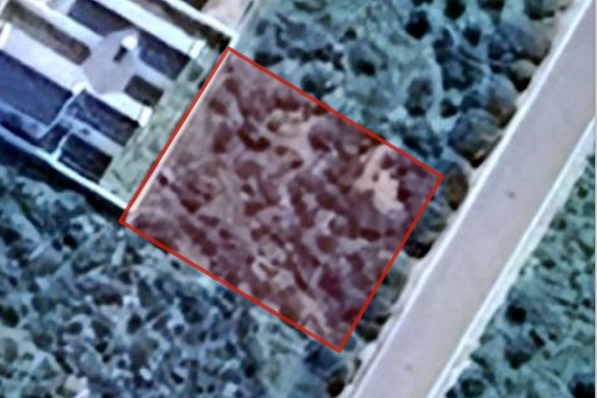 An aerial view of a vacant residential plot, highlighted in red, showing its location relative to the surrounding landscape and nearby road.