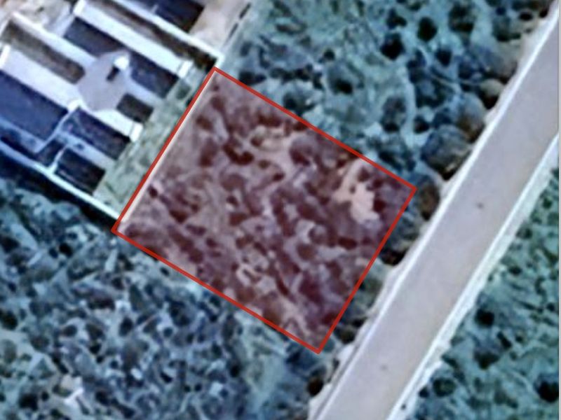 An aerial view of a vacant residential plot, highlighted in red, showing its location relative to the surrounding landscape and nearby road.