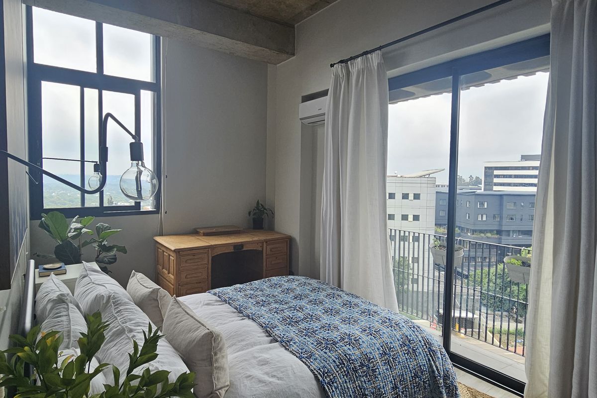 A bright bedroom featuring large windows, a sliding door leading to a balcony, and exposed concrete ceiling details.