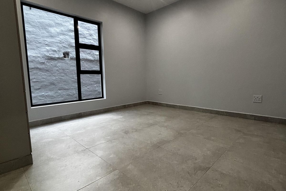 A clean, empty bedroom featuring neutral large-format floor tiles, recessed ceiling lighting, and a modern black-framed window overlooking a textured exterior wall.