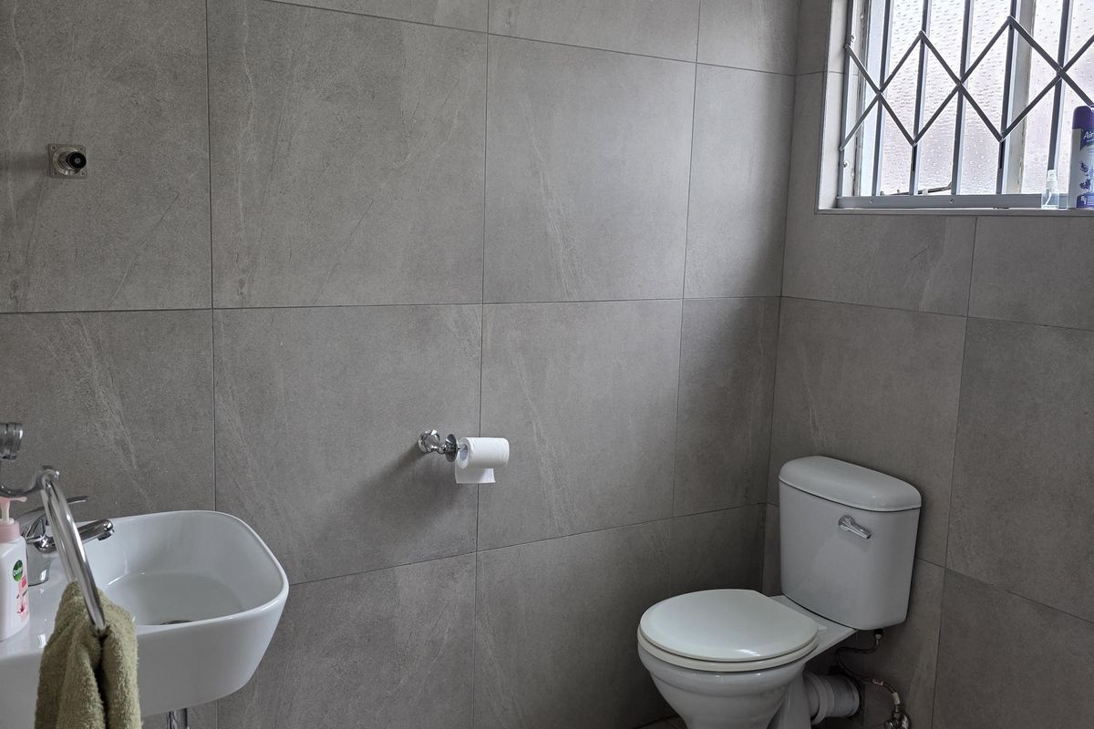 A modern bathroom featuring large-format grey wall and floor tiles, a wall-mounted basin, and a standard toilet with a security-grille window.