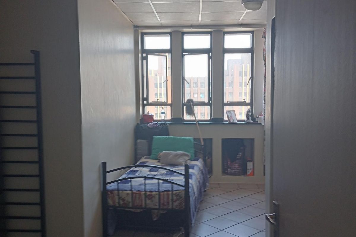 A compact bedroom featuring tiled flooring, a drop ceiling, and large windows providing natural light and an urban view.