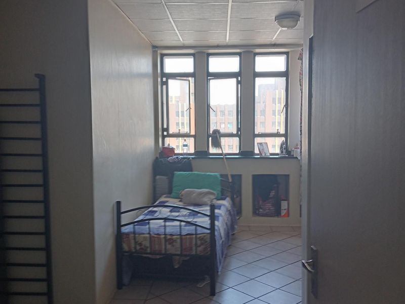 A compact bedroom featuring tiled flooring, a drop ceiling, and large windows providing natural light and an urban view.