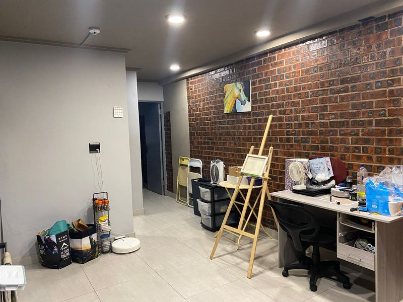 A living area featuring a prominent exposed brick feature wall, recessed ceiling lighting, and neutral floor tiling, leading into a hallway.
