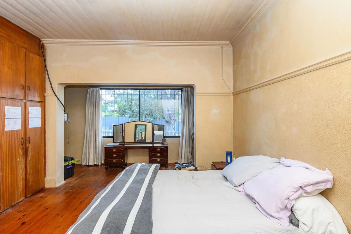 Spacious bedroom featuring original timber flooring, built-in wooden cupboards, and large windows with security bars, offering a classic layout with potential for modernization.