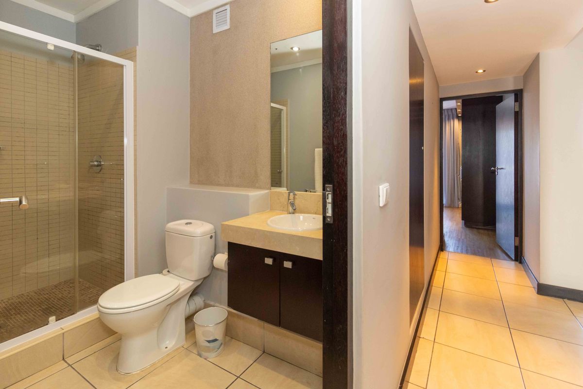 A modern bathroom featuring a glass-enclosed shower, a vanity with a basin, and a toilet, connected to a tiled hallway.