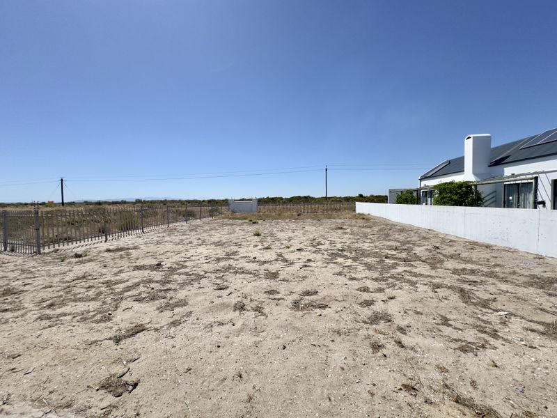 A vacant residential plot featuring sandy terrain, enclosed by perimeter fencing and adjacent to an existing property with a boundary wall.