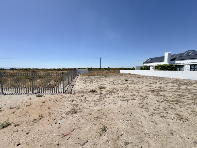 A view of a vacant residential plot featuring sandy ground, perimeter metal fencing, and a boundary wall adjacent to an existing property.