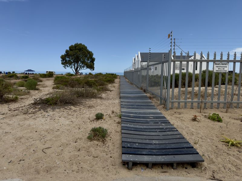 A sandy path featuring a wooden boardwalk leading towards the coast, bordered by a secure metal perimeter fence and natural coastal vegetation.