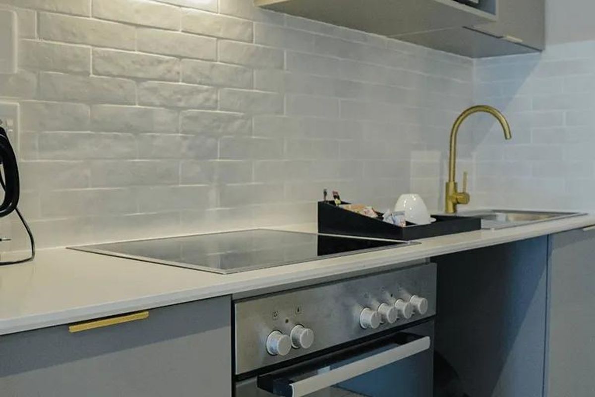 Modern kitchen featuring grey cabinetry, white stone countertops, a subway tile backsplash, integrated electric cooktop, oven, and a contemporary brass faucet.