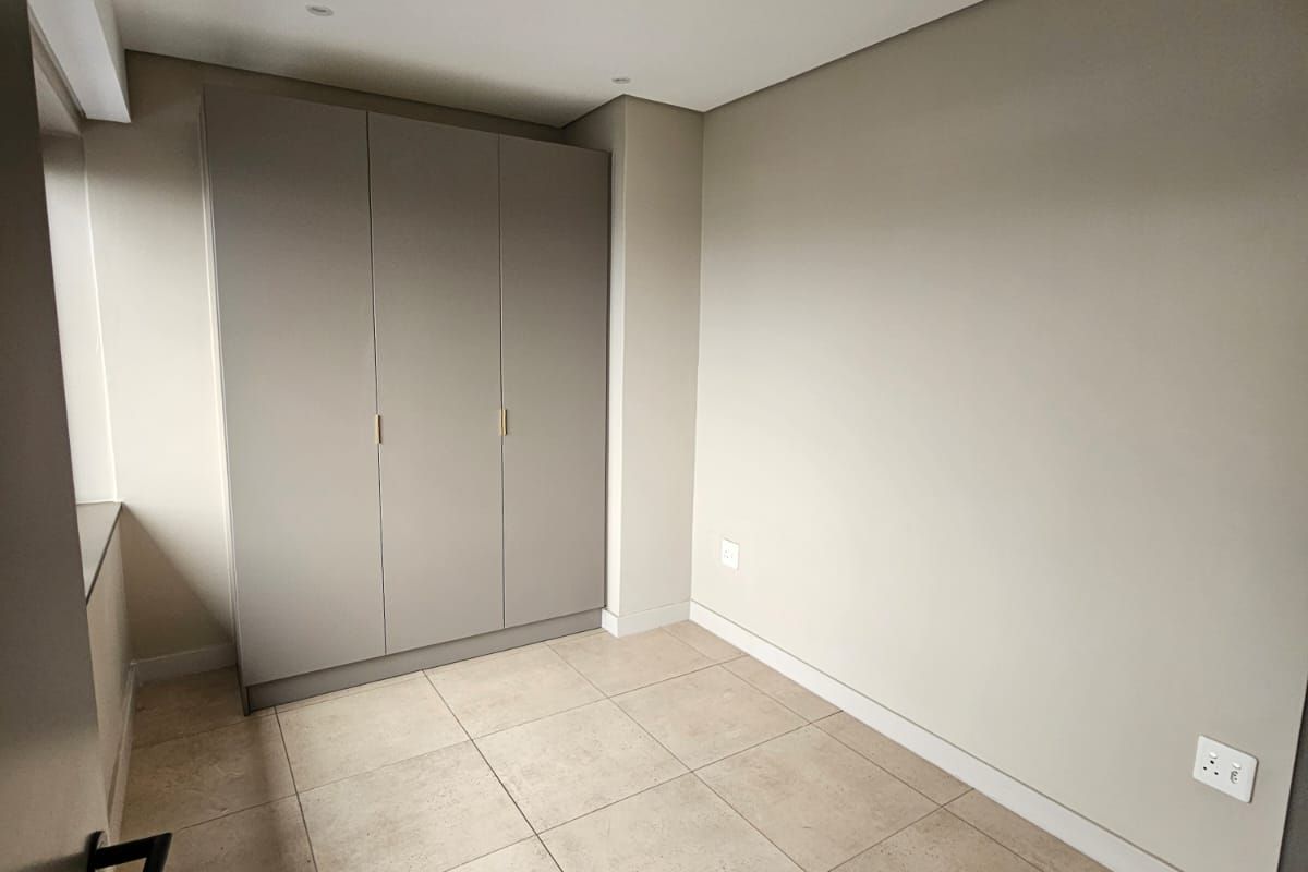 A clean, modern bedroom featuring neutral wall tones, large format floor tiles, and built-in cabinetry with minimalist hardware.