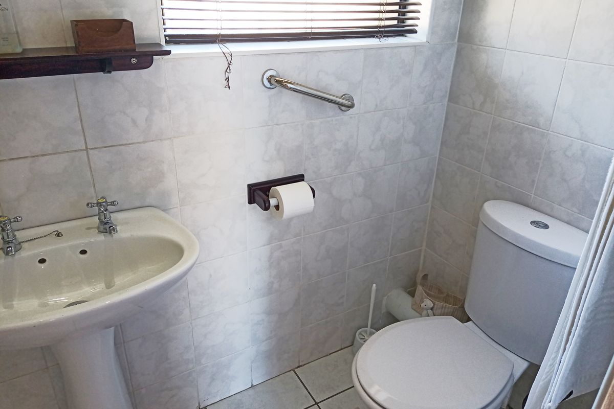 A functional bathroom featuring a pedestal basin, toilet, and tiled finishes, equipped with a safety grab rail and window blinds.