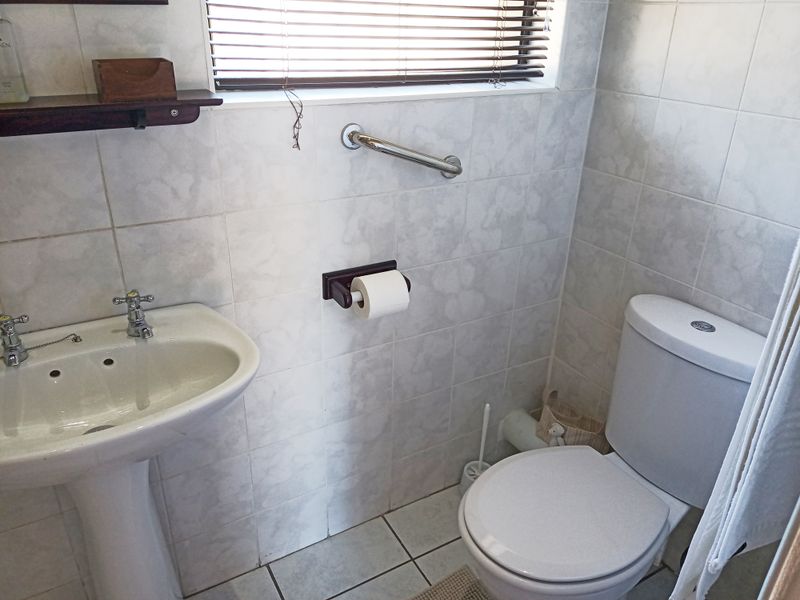 A functional bathroom featuring a pedestal basin, toilet, and tiled finishes, equipped with a safety grab rail and window blinds.