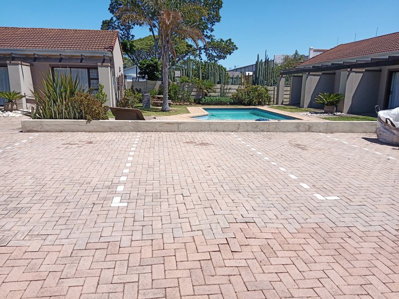 Rear exterior view featuring a paved parking area, a swimming pool, and a pergola structure set within a landscaped garden.