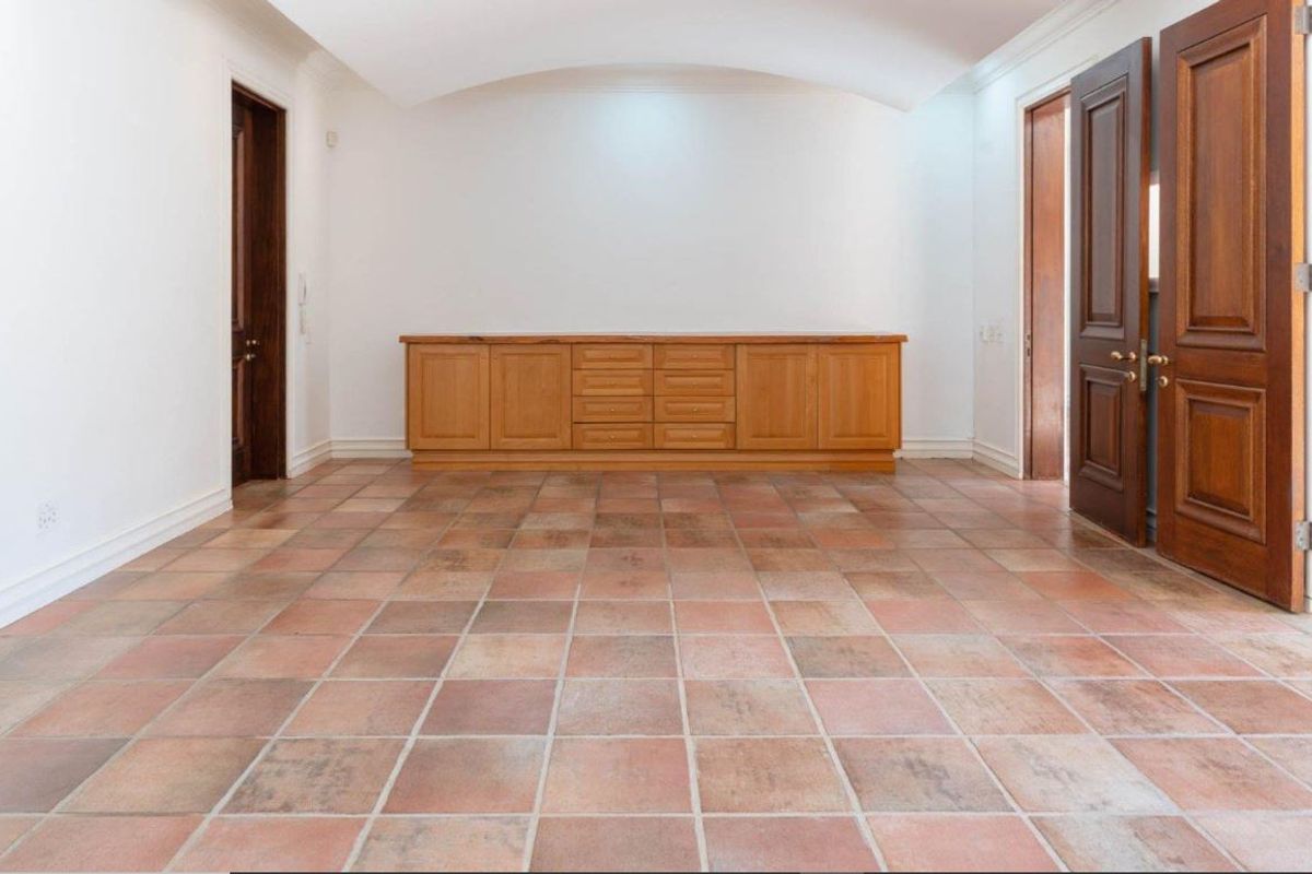 A spacious, empty living area featuring terracotta floor tiles, a built-in wooden cabinet, and arched ceiling details with solid wood doors.