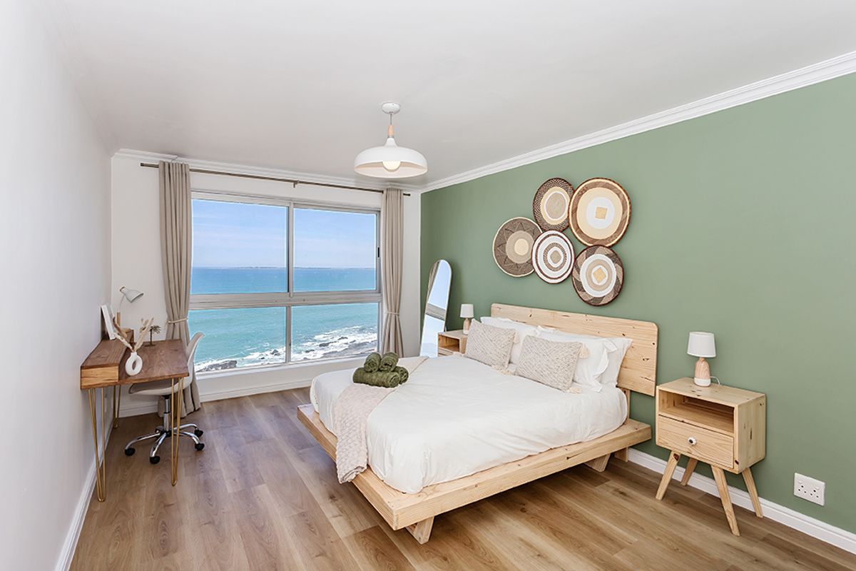 A bright bedroom featuring wood-look flooring, a sage green accent wall, and large windows offering expansive ocean views.