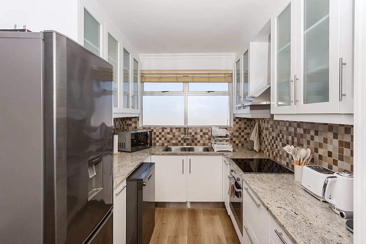 A modern galley-style kitchen featuring granite countertops, white cabinetry with glass inserts, a built-in oven, hob, extractor fan, and double stainless steel sink.