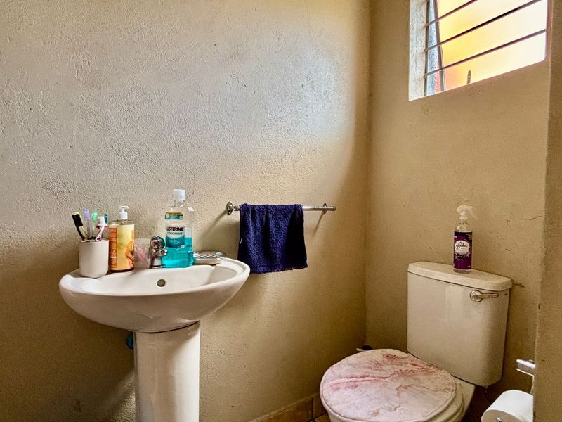 A compact bathroom featuring a pedestal basin, standard toilet, and a small window with security bars, requiring cosmetic updates to improve its overall condition.