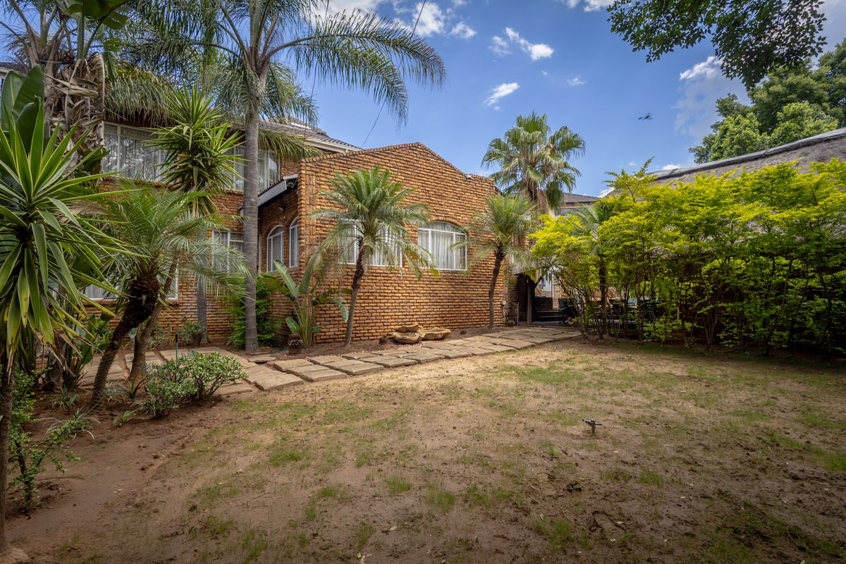 A rear view of the property featuring a classic face-brick facade, established garden with mature palm trees, and a paved stone walkway.