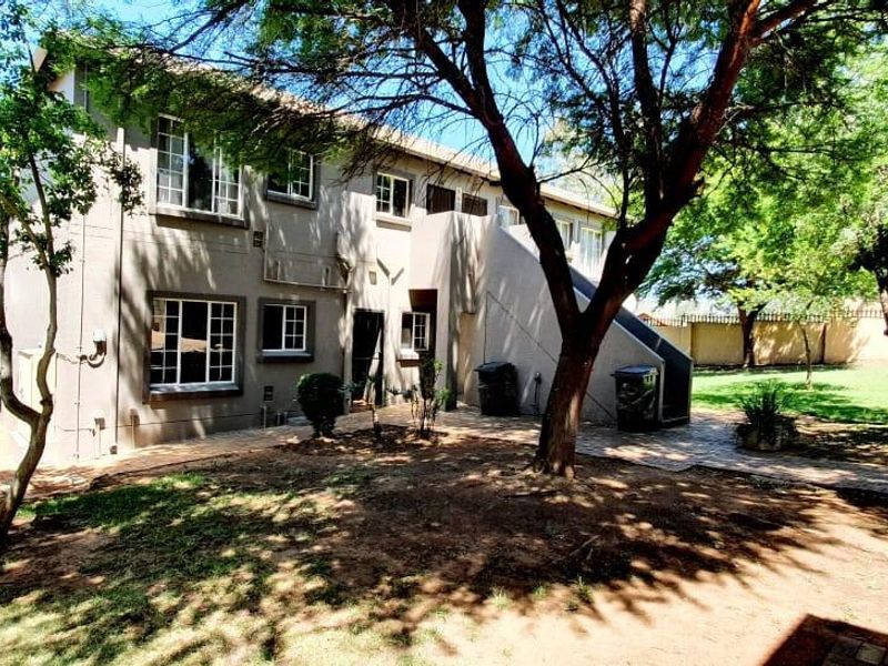 Rear view of the apartment building featuring a paved walkway, external staircase, and surrounding garden area with mature trees.