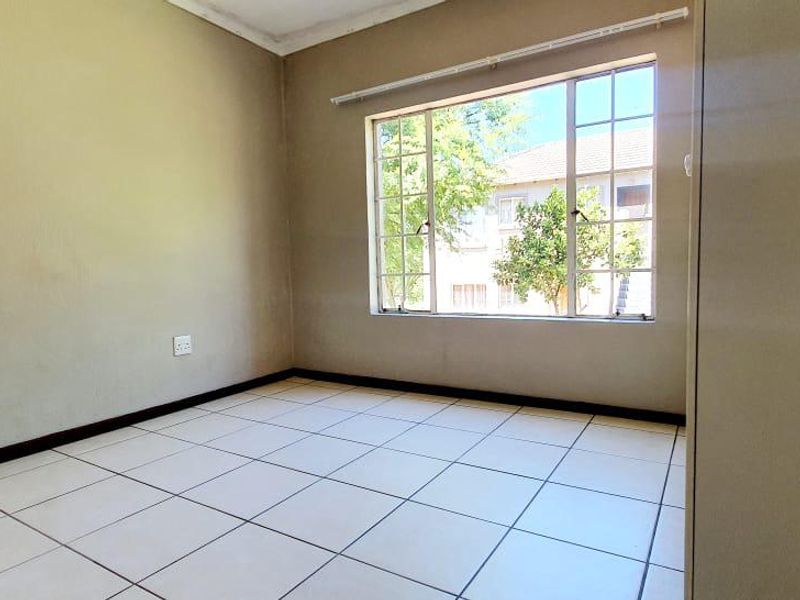 A bright, empty bedroom featuring neutral tiled flooring, a large window providing natural light, and a built-in cupboard.