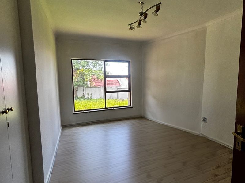 A bright, empty bedroom featuring modern laminate flooring, built-in cupboards, and a large window providing natural light.