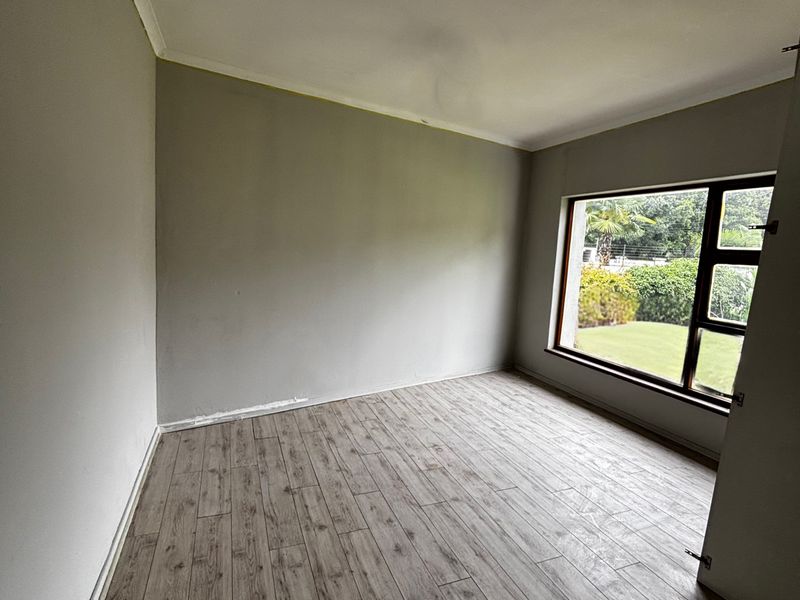 A bright, empty bedroom featuring modern wood-look laminate flooring, a large window overlooking the garden, and neutral wall tones.