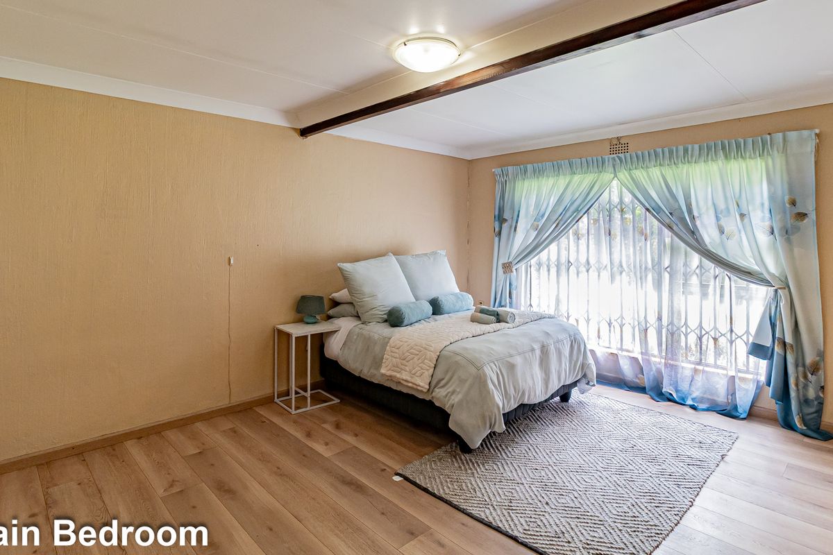 A spacious bedroom featuring light wood-look flooring, a central ceiling light, and a large window fitted with security bars and curtains.