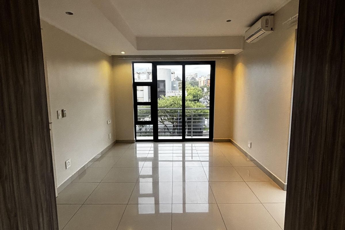 A bright, empty living area featuring polished floor tiles, recessed ceiling lighting, a wall-mounted air conditioning unit, and large glass sliding doors leading outside.