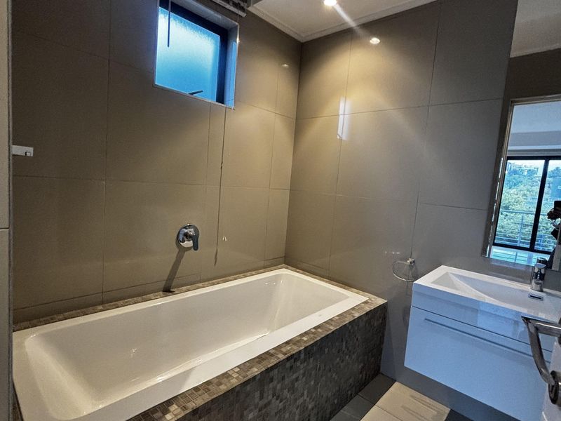 Modern bathroom featuring a built-in bathtub with mosaic tile surround, wall-mounted vanity, and large format neutral wall and floor tiles.