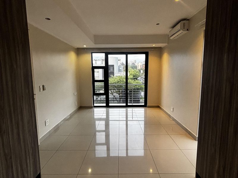 A bright, empty living area featuring polished floor tiles, recessed ceiling lighting, a wall-mounted air conditioning unit, and large glass sliding doors leading outside.