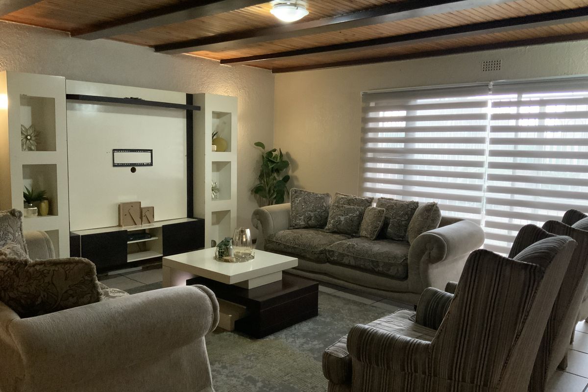 A comfortable living room featuring a built-in media unit, warm timber beam ceiling, and large windows with modern roller blinds.
