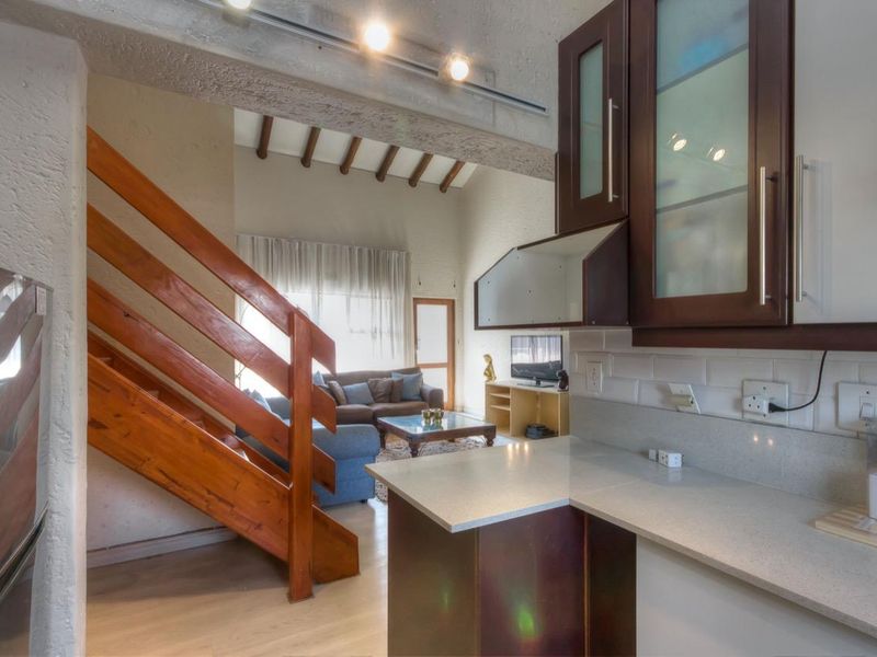 Open-plan kitchen featuring dark cabinetry and stone countertops, seamlessly flowing into a living area with a wooden staircase and high vaulted ceilings.