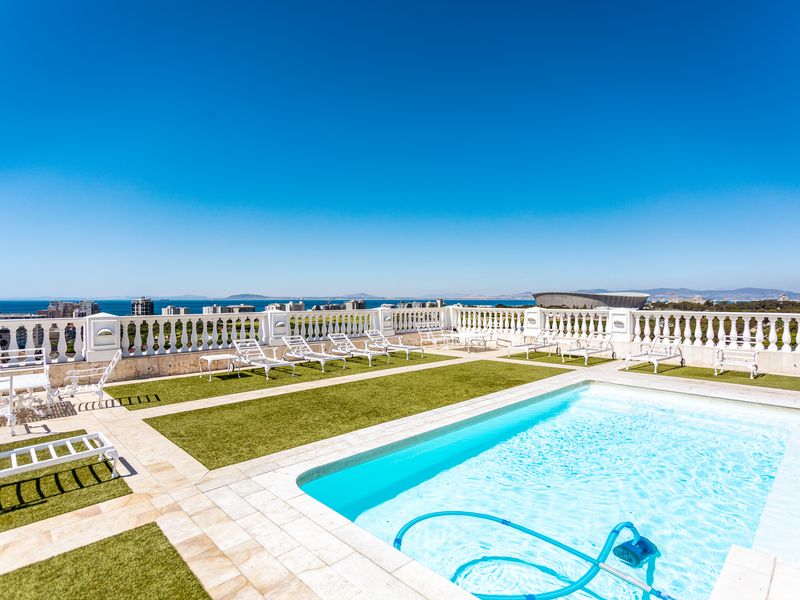 Spacious rooftop pool area featuring a swimming pool, stone paving, and decorative balustrades with panoramic coastal and city views.