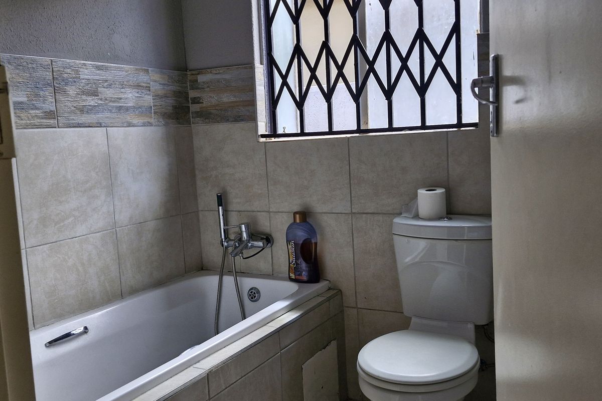 A functional bathroom featuring a built-in bathtub with shower mixer, a standard toilet, and tiled wall finishes with a security gate on the window.