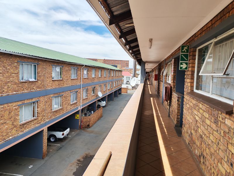 An exterior view of a residential apartment complex balcony featuring brick construction, tiled flooring, and access to covered parking areas below.